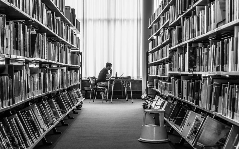 Young man studying in library, black and white image with book shelfs on either side.